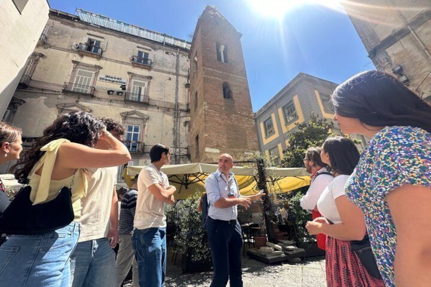 Un gruppo di persone in piedi all'aperto, in una zona storica con un'alta torre e edifici sullo sfondo. 3