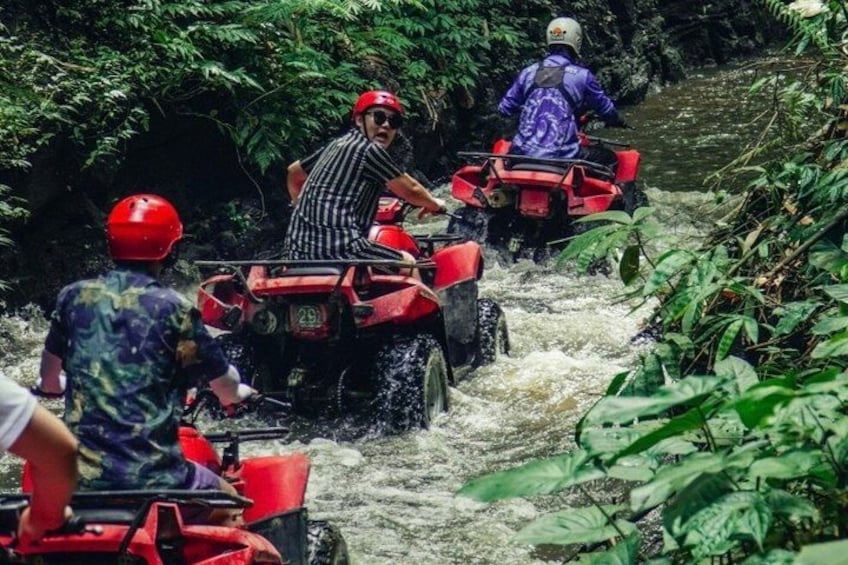People riding red ATVs through a narrow, water-filled river surrounded by dense greenery. 2