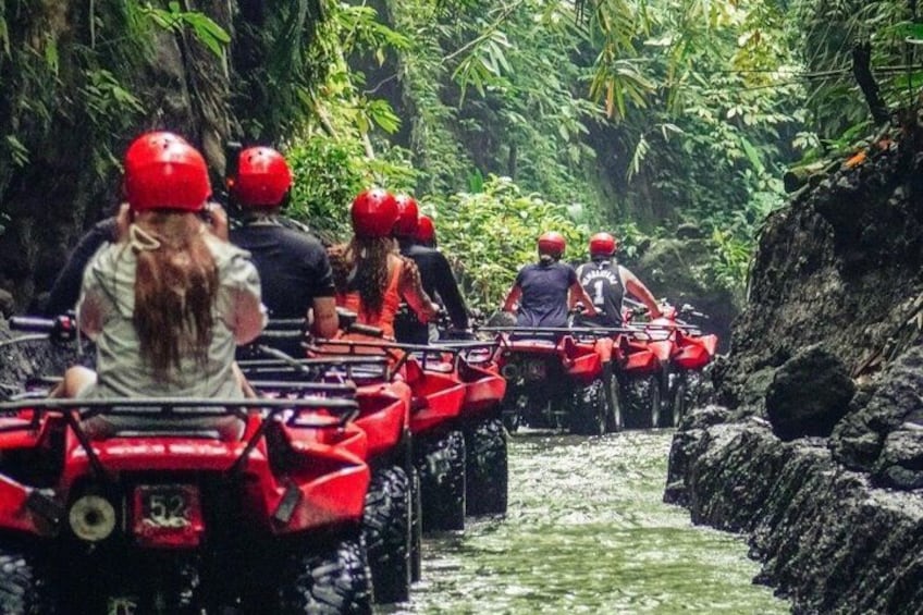 People riding red ATV's through a narrow water channel surrounded by dense greenery. 5