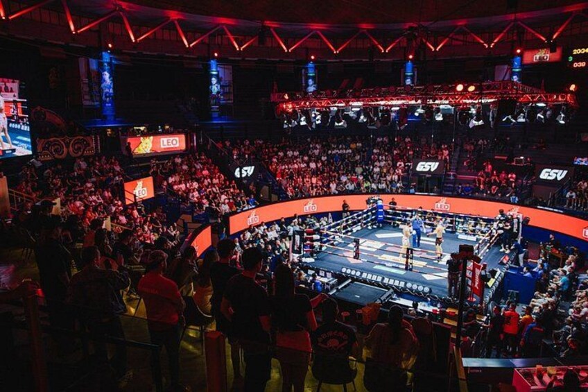 A crowded indoor arena with a boxing ring at the center, surrounded by spectators. 5