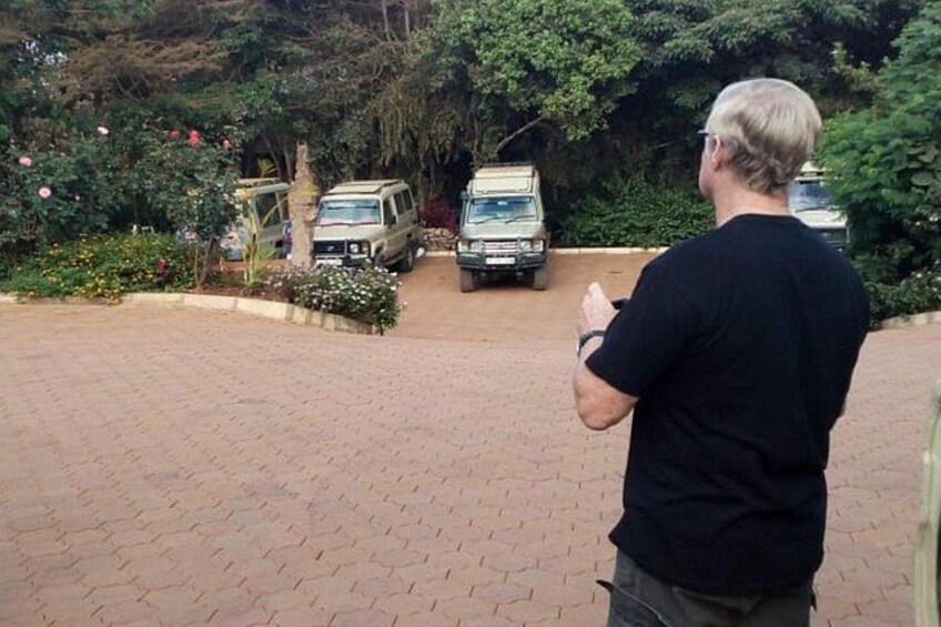 A paved driveway with two vehicles parked, surrounded by lush greenery and flowering plants. 2