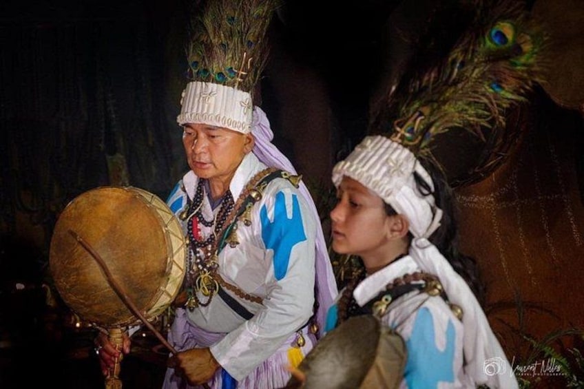 Två personer klädda i traditionella dräkter, med påfågelfjädrar, varav en spelar på en trumma. 1