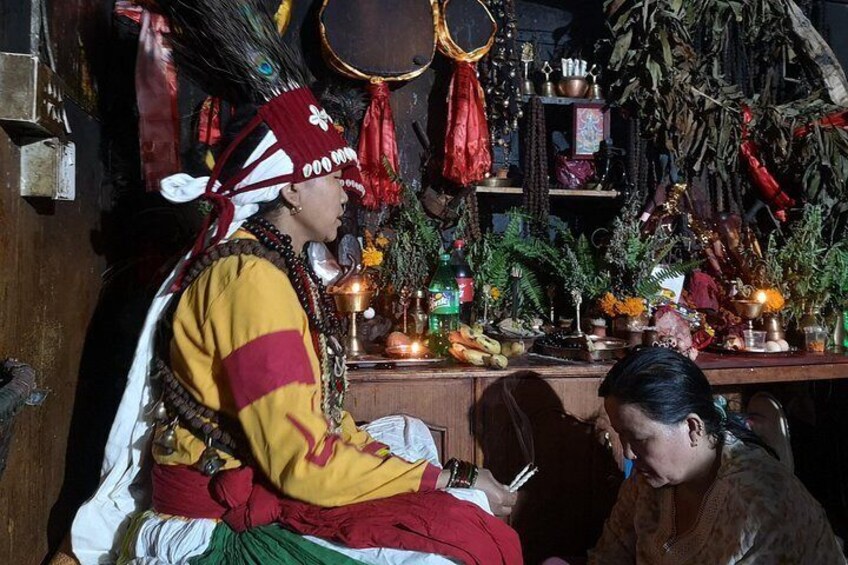 Une personne vêtue d’un costume traditionnel est assise dans une pièce où l’on peut voir différentes étagères remplies de divers objets, notamment des plantes et des récipients. 2