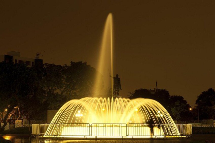 Una fontana illuminata di notte, con getti d'acqua e una statua, circondata da un recinto e alberi. 3