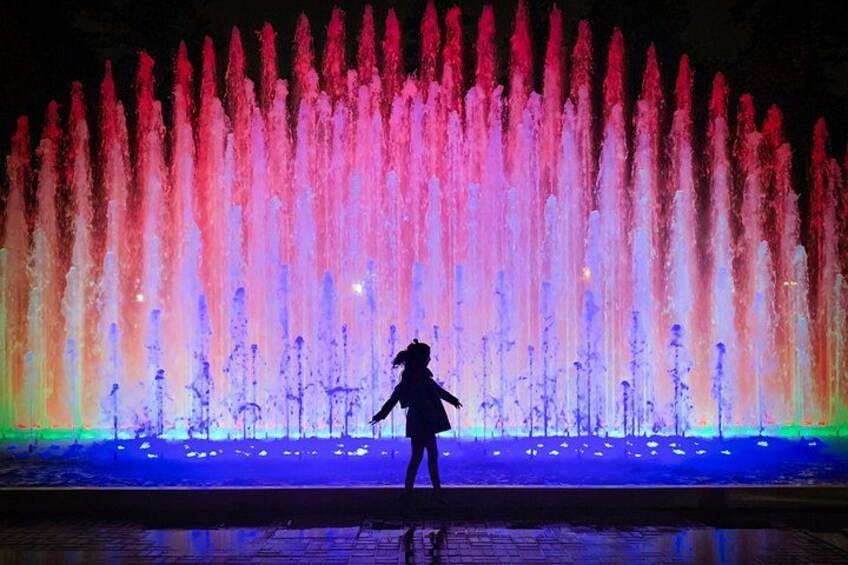 Una persona in piedi di fronte a una fontana colorata illuminata di notte. 1