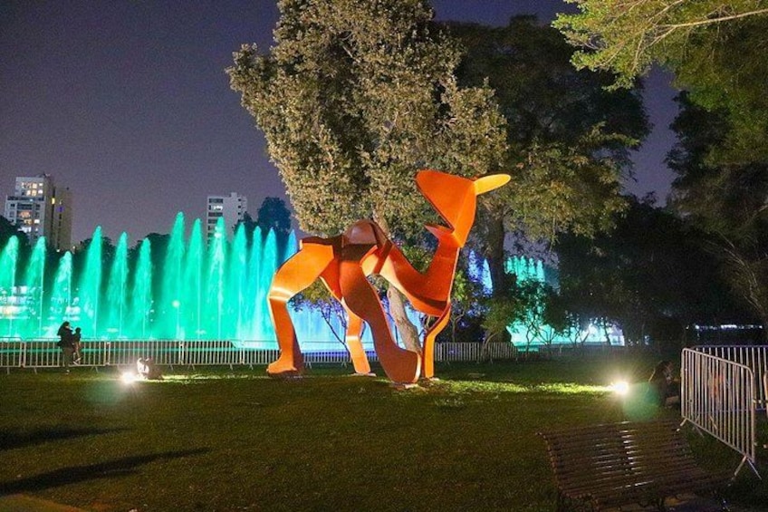 A park at night with a large illuminated sculpture and a fountain in the background. 3