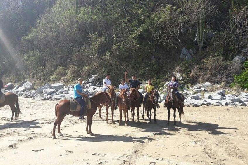 En grupp människor som rider på hästar längs en sandstrand, med klippor och träd i bakgrunden. 5