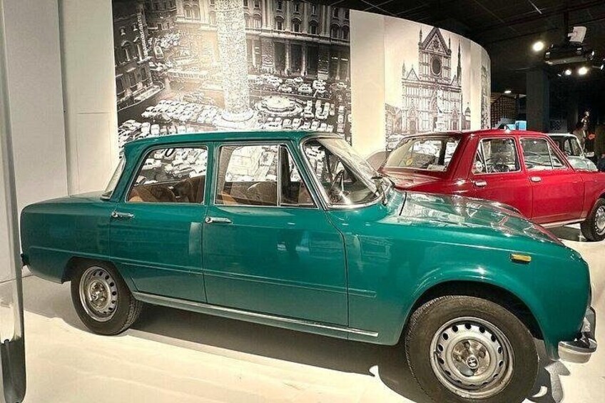 Une voiture ancienne de couleur turquoise est exposée devant une fresque représentant un paysage urbain. 5