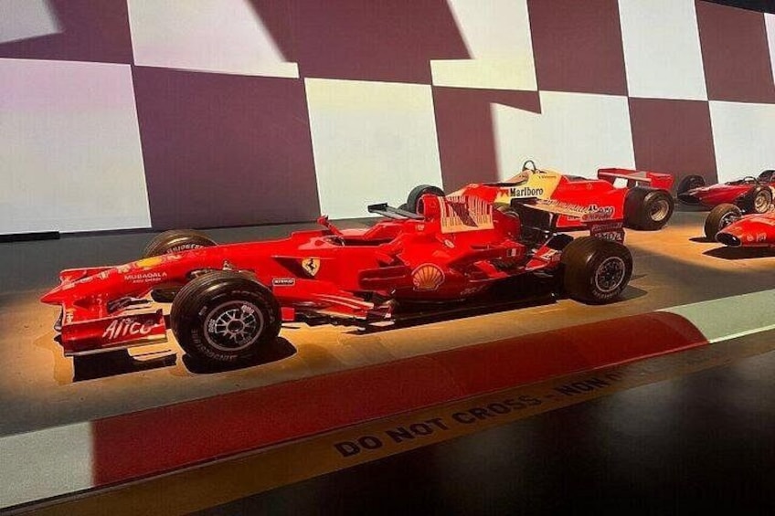 Une voiture de Formule 1 rouge, ornée des logos de ses sponsors, est exposée sur une plateforme dont le sol présente un motif à carreaux. 3