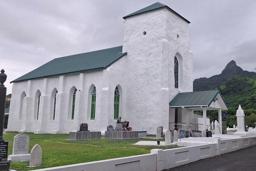 En vit kyrka med ett grönt tak, en liten sidobyggnad och en kyrkogård med gravstenar. 4
