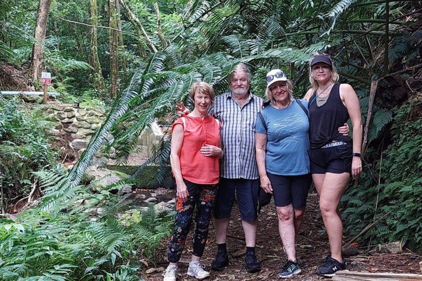 En grupp människor står i ett skogsområde med tät vegetation, och i bakgrunden syns en stenmur. 2