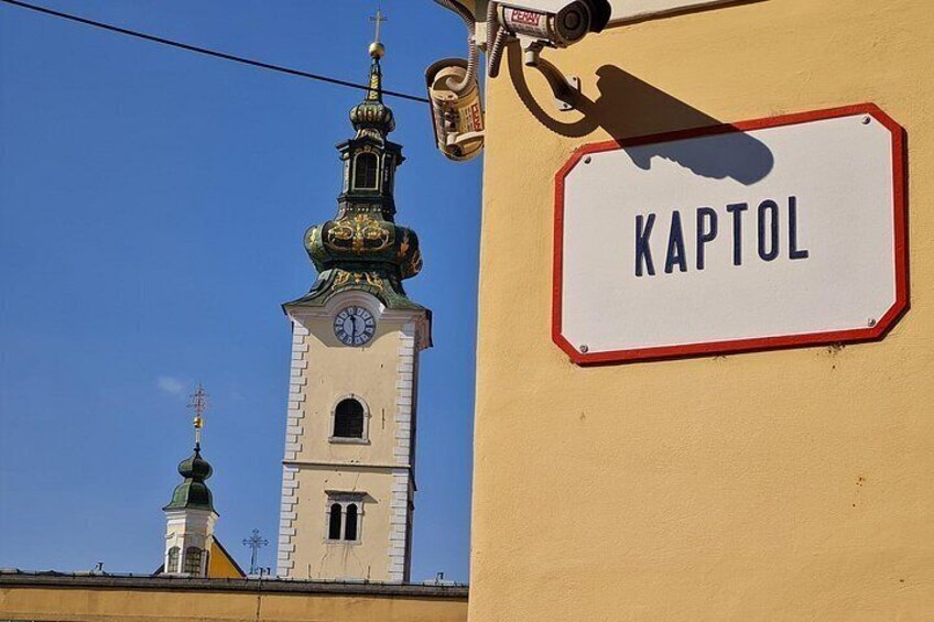 Una torre de iglesia con un reloj y un letrero que dice 'KAPTOL' sobre un cielo azul despejado. 2