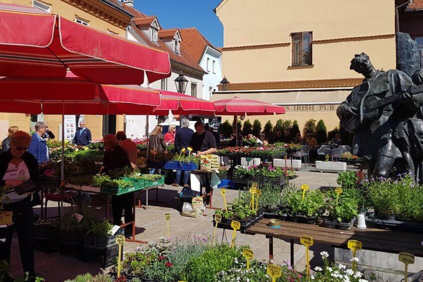 Un mercado al aire libre concurrido, con sombrillas rojas, gente comprando y una estatua al frente. 4