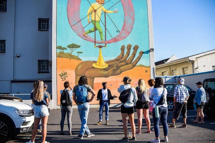 Un mural que muestra a un hombre equilibrándose en un poste, con una mano sosteniéndolo, rodeado de varias personas. 5