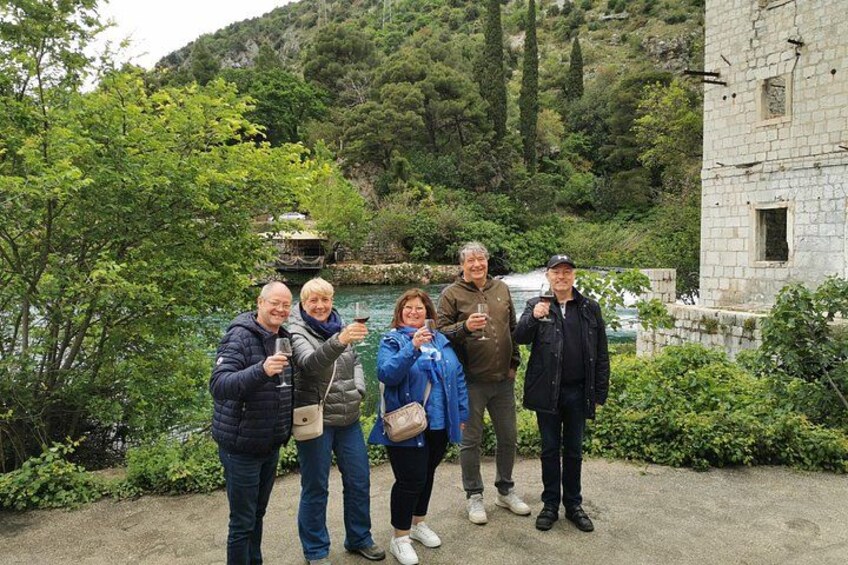 A group of people standing by a body of water with trees and a stone building in the background. 5