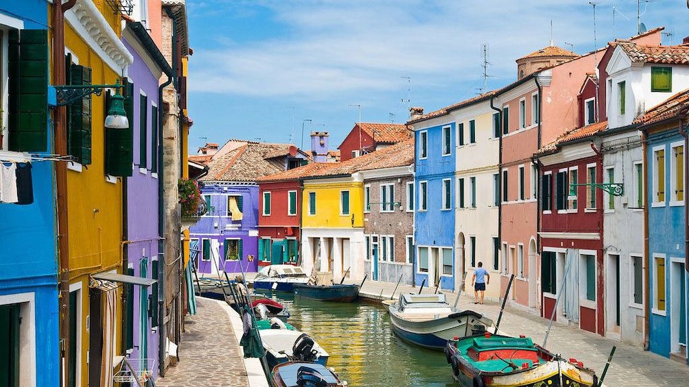Un canal étroit bordé de bâtiments aux couleurs vives, des bateaux amarrés le long du rivage et une personne qui marche au loin. 1