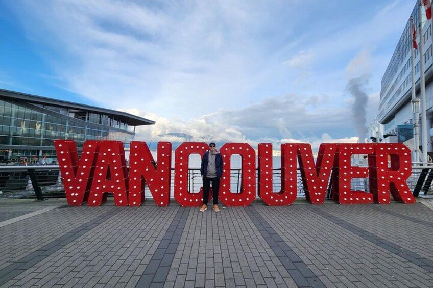 Grandes letras iluminadas que deletrean 'VANCOUVER' sobre una superficie pavimentada, con un edificio moderno de fondo. 2