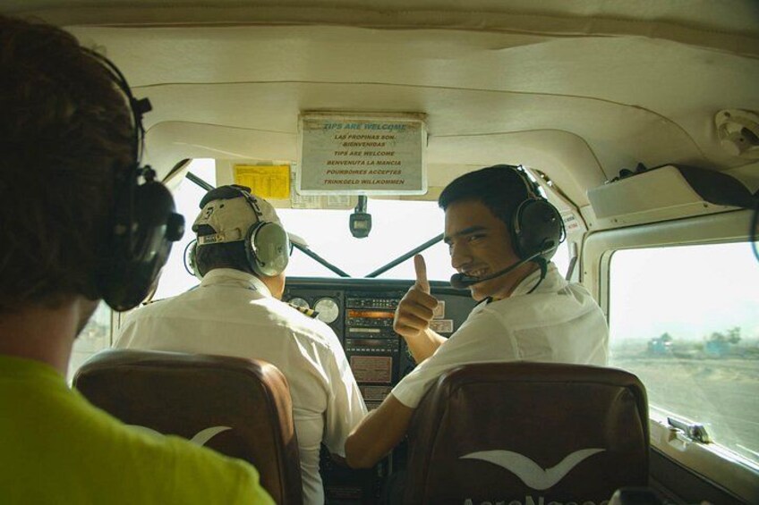 Cabina de vuelo con dos pilotos, uno levantando el pulgar, y un cartel en la pared. 2