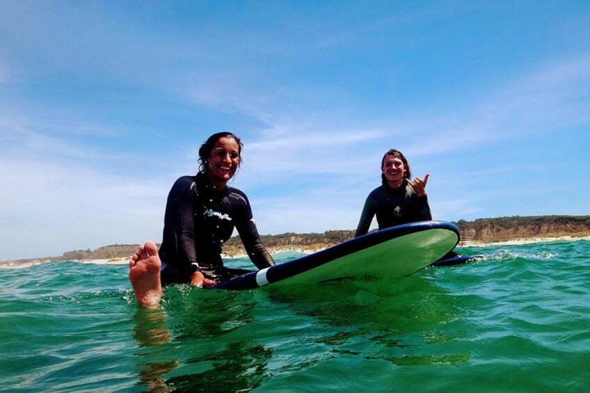 Two people in wetsuits on a surfboard in the ocean with a beach in the background. 1