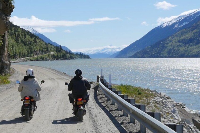 Dos motociclistas en un camino de tierra al lado de un lago con montañas al fondo. 2