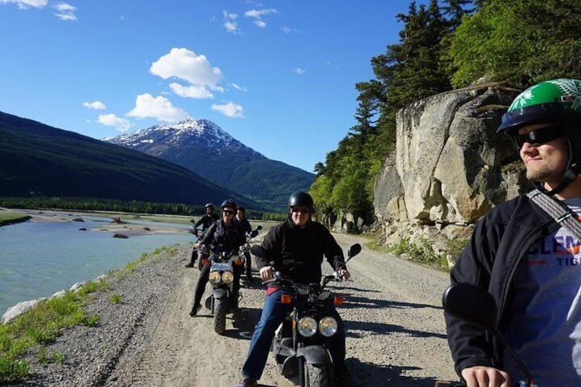 Un grupo de motociclistas en un camino de grava, con un río y montañas al fondo. 2