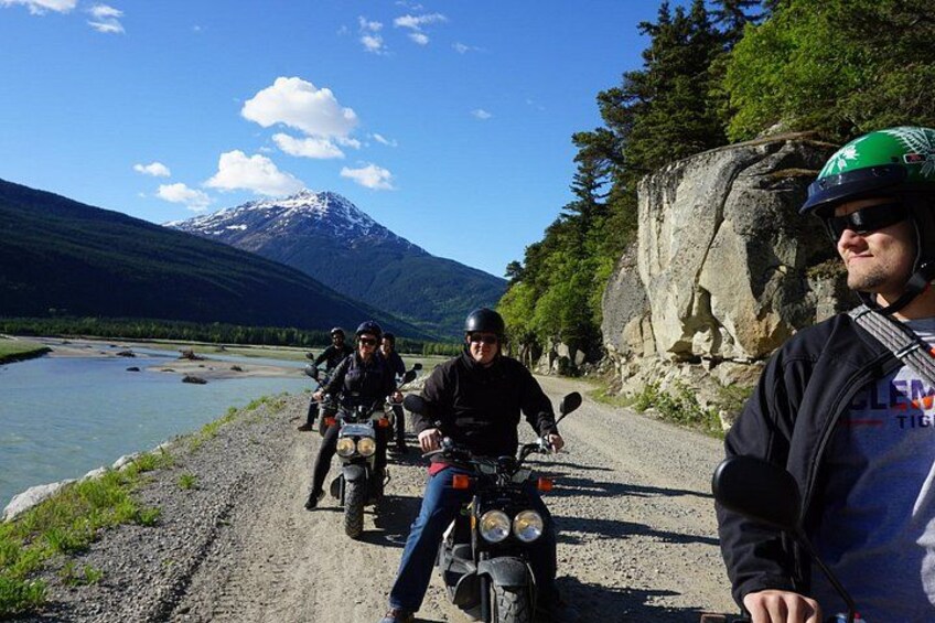 En grupp motorcyklister på en grusväg med en flod och berg i bakgrunden. 3