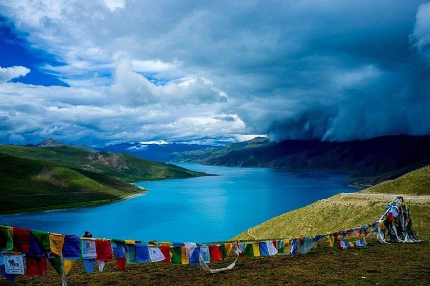 A lake surrounded by mountains, a person standing on a hill, and colorful prayer flags. 1