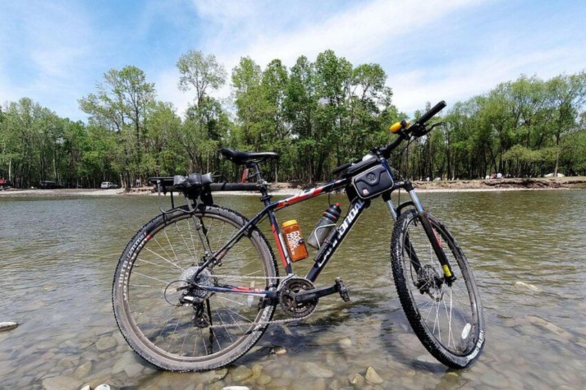 Una bicicleta de montaña con una botella de agua y guardabarros, estacionada a orillas de un río tranquilo con árboles al fondo. 4