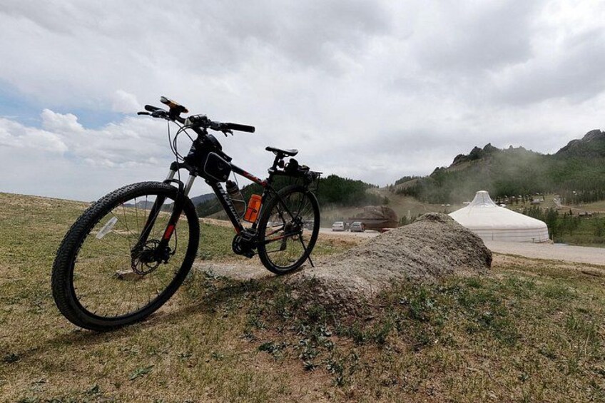 Una bicicleta de montaña estacionada en un campo cubierto de hierba, con una roca grande, una tienda de campaña y un cielo nublado al fondo. 2