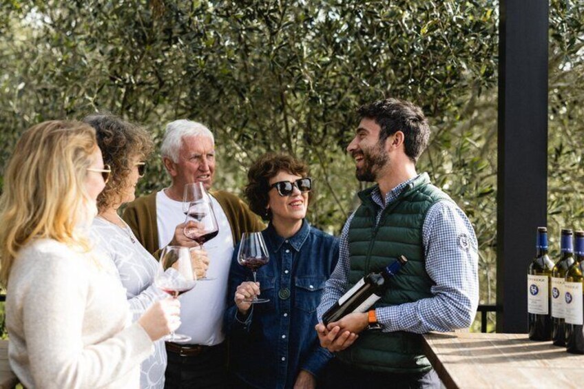 A group of people standing outdoors, some holding wine glasses, with a row of wine bottles in the background. 1