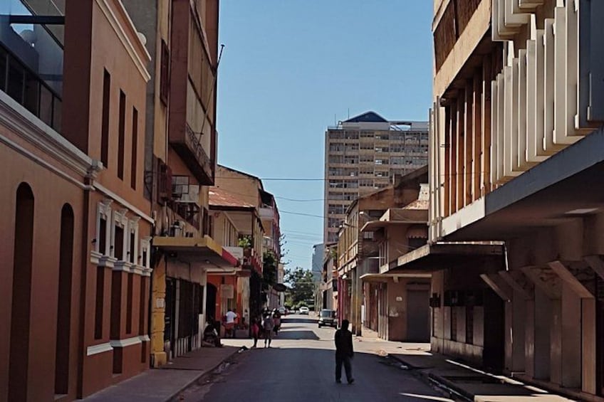 Una calle con edificios, una persona caminando y un coche estacionado al lado. 2