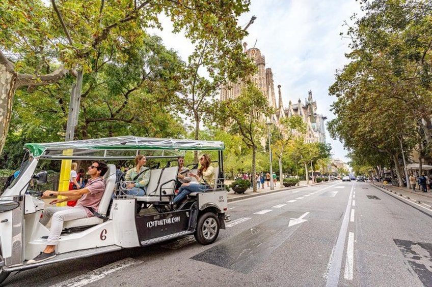 En vit turistbuss med passagerare kör på en stadsgata som kantas av träd och byggnader. 2