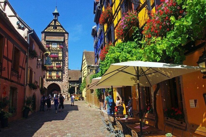Eine Kopfsteinpflasterstraße mit Gebäuden, einem Turm und Passanten. 5