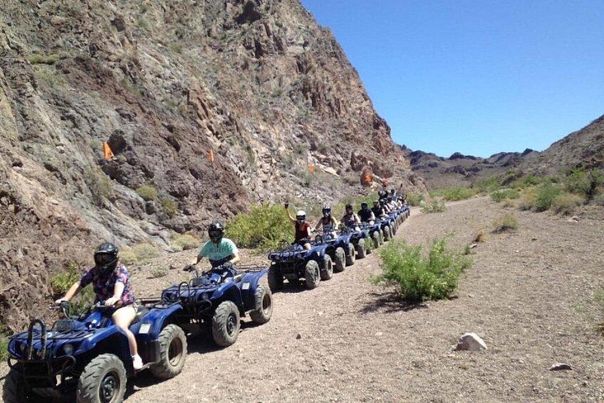 A group of people riding ATVs on a dirt path in a desert landscape with rocky hills. 2