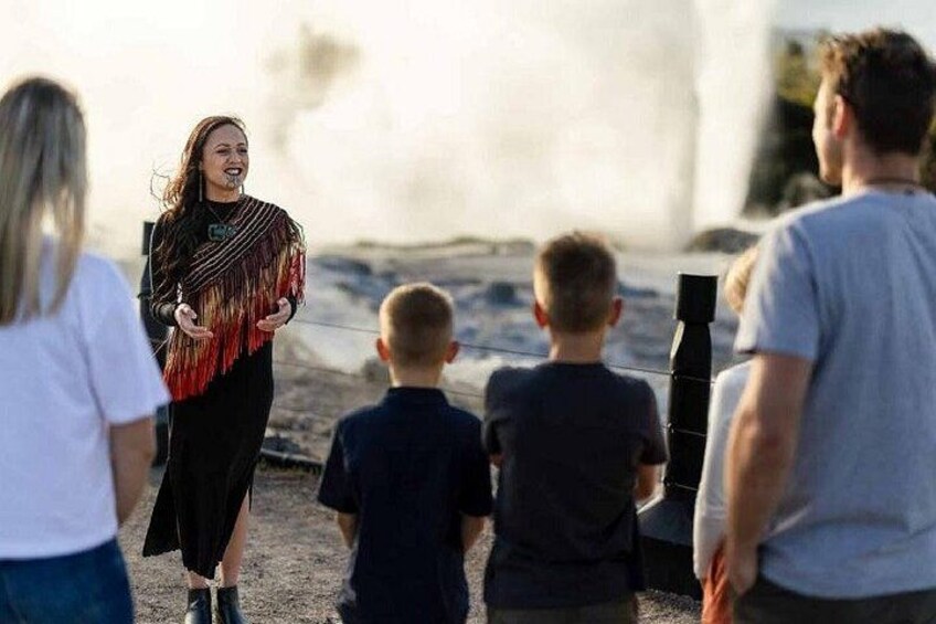 A person in a black dress with a fringe shawl, standing on a sandy beach with a group of people, including children, looking at something in the distance. 5