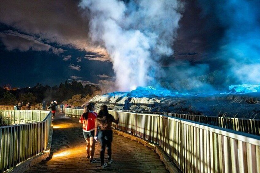 A wooden boardwalk with people walking, a geyser emitting steam, and a snowy landscape in the background. 1