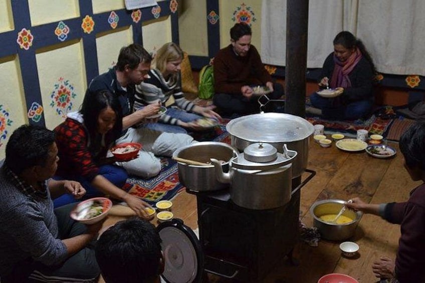 A group of people sitting on the floor, eating from bowls and plates, with large pots and a stove in the center. 2