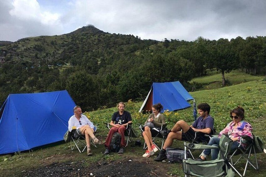 A group of people sitting on chairs and camping tents in a grassy field with hills in the background. 4