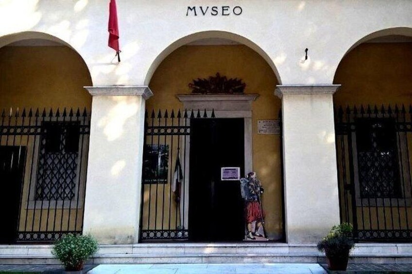 Un bâtiment sur lequel on peut lire l’inscription « MVSEO » au-dessus de l’entrée, une statue représentant une personne et un drapeau rouge. 5