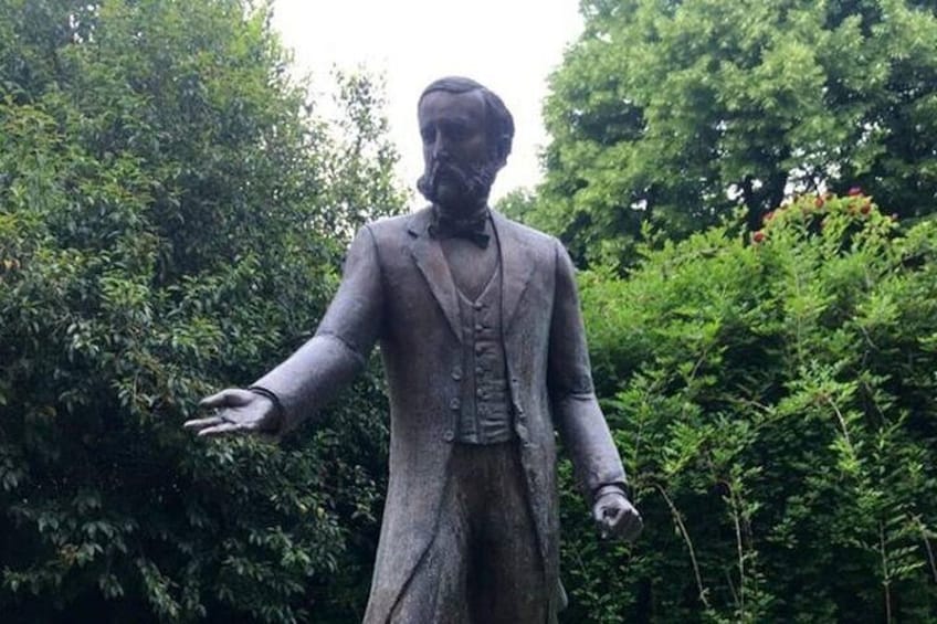 Une statue représentant un homme en costume, les bras tendus, se dressant sur un fond de feuillage dense. 4