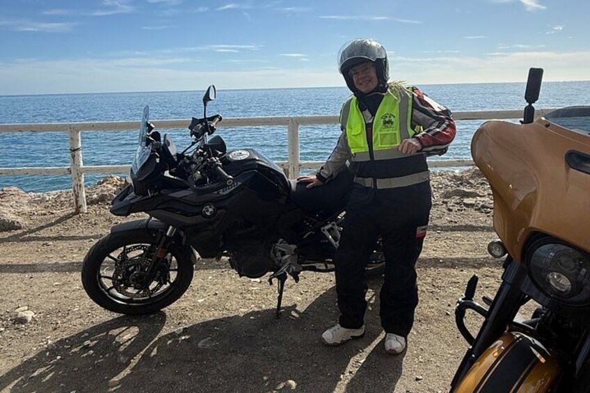 A person standing next to a motorcycle by a coastal road with a fence and a yellow vehicle in the background. 2