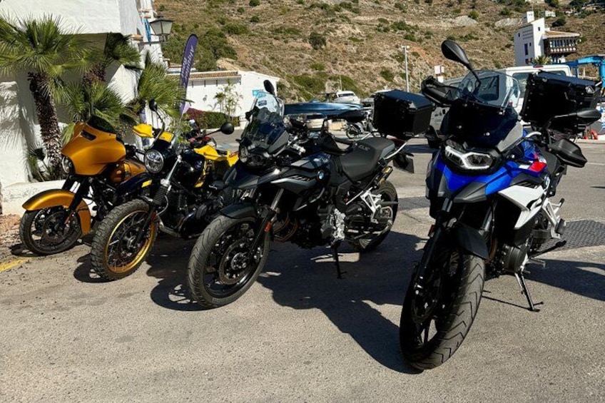 A row of motorcycles parked on a paved surface with a building and palm trees in the background. 1