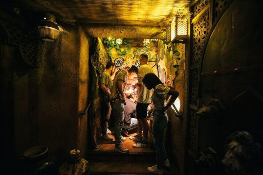 Un estrecho pasaje con gente reunida alrededor de una figura central, posiblemente un artista o guía, en un entorno rústico y tenuemente iluminado con detalles decorativos. 1
