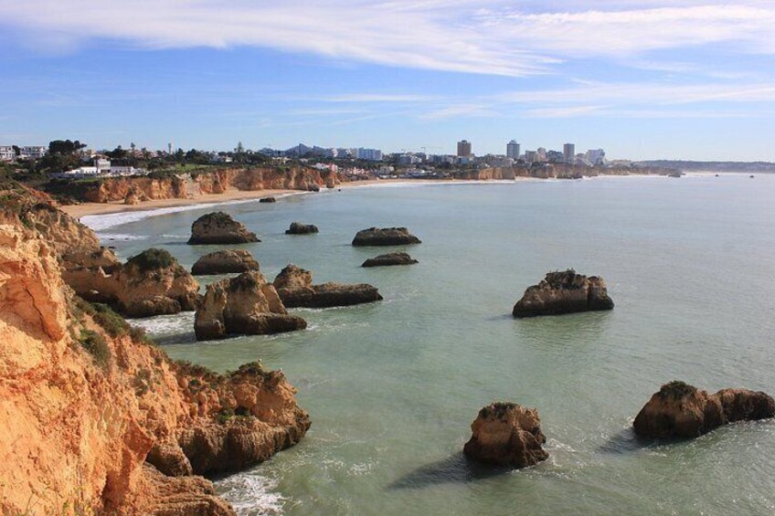 Un paisaje costero con afloramientos rocosos, una playa arenosa y el horizonte de una ciudad a lo lejos. 4