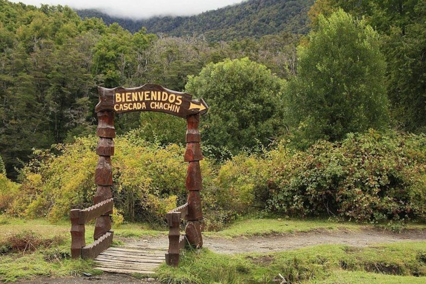 A rustic wooden signpost with the words 'BIENVENIDOS CASCADA CHACHIN' and a wooden footpath leading through a forested area. 3