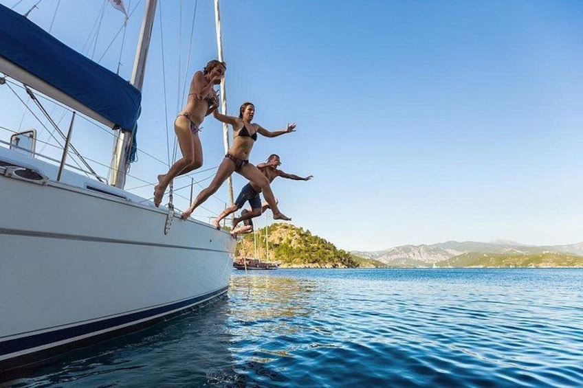 People jumping off a sailboat into a body of water with a distant shoreline and mountains in the background. 1