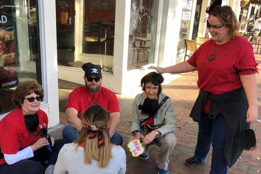 A group of people sitting on the sidewalk, some wearing red shirts, with a storefront in the background. 5