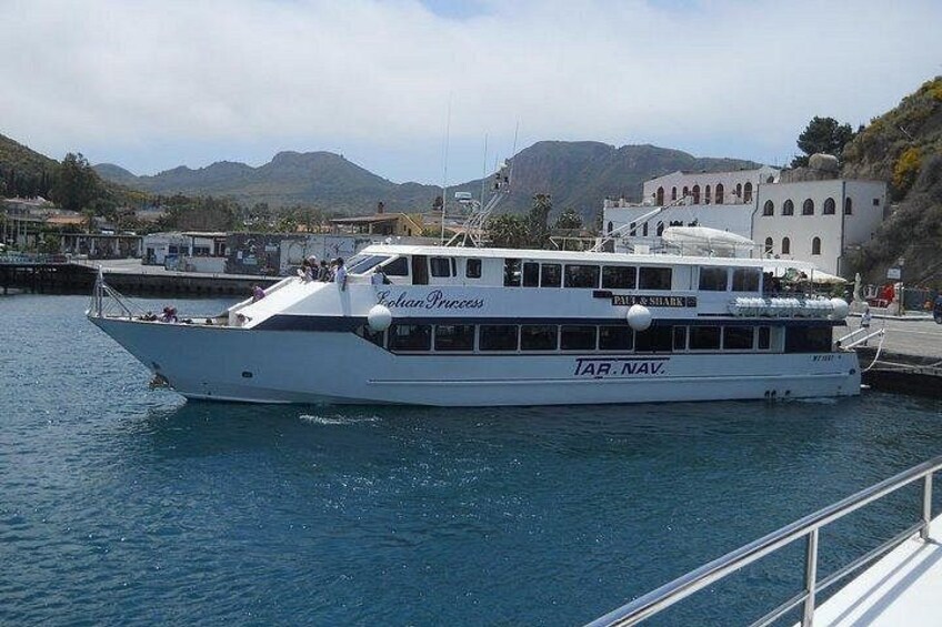 A white boat named 'Tar. Nav.' docked at a pier with a mountainous landscape in the background. 3