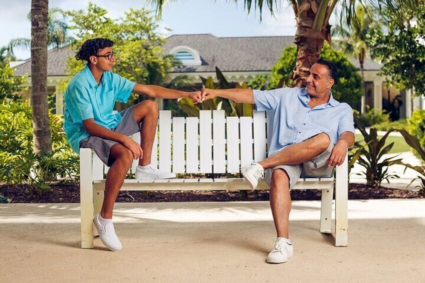 Two people sitting on a white bench outdoors with greenery and a building in the background. 1
