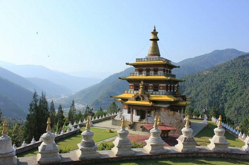 A multi-tiered pagoda with a golden roof, surrounded by a landscaped garden with white stupas, set against a mountainous backdrop. 1